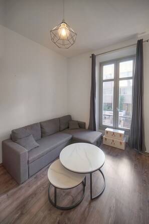 Ein modernes Wohnzimmer mit einem grauen Ecksofa, zwei runden Tischen mit Marmorplatten, einer Hängeleuchte und einem Fenster mit Vorhängen.