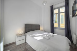 Une chambre à coucher comprenant un lit, des lampes de chevet, une table de chevet, une fenêtre avec des rideaux et un luminaire au plafond.