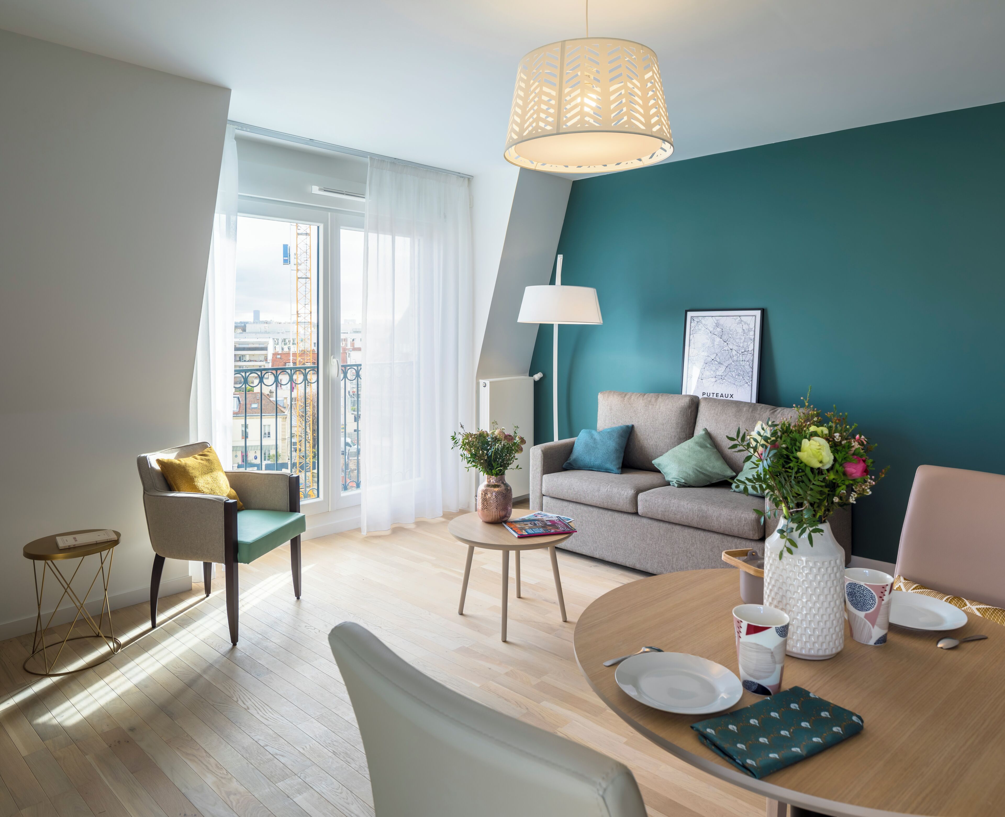 Un salon moderne avec un mur d’accent de couleur turquoise, un canapé agrémenté de coussins, une table à manger avec de l’argenterie et un vase de fleurs, et une vue sur la ville par une grande fenêtre.