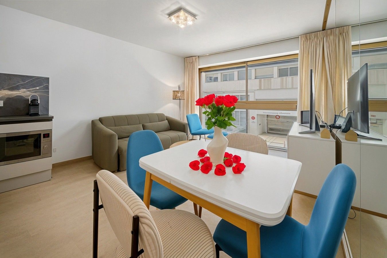 A modern hotel room with a dining area, a sofa, a table with a vase of red flowers, and a kitchen area with a microwave.