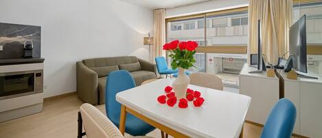 A modern hotel room with a dining area, a sofa, a table with a vase of red flowers, and a kitchen area with a microwave.