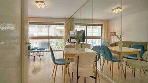 Ein modernes Wohnzimmer mit Glastrennwand, einem Esstisch, blauen Stühlen, einer Couch, einem Fernseher und einer Pflanze.