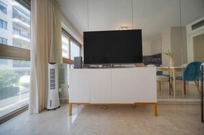 A modern living room with a flat-screen TV on a white cabinet, a dining area with a table and chairs, and a large window with curtains. - One-bedroom apartment in Nice, France (Niza)