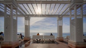 Ein überdachter Außenbereich mit Bänken, einer Pergola und Blick aufs Meer.