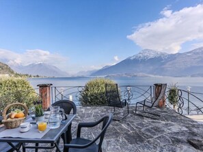 Uma mesa com cadeiras e uma cesta de frutas, com vista para um lago e montanhas ao fundo.