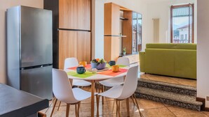 A modern kitchen with a built-in refrigerator, white chairs, a dining table with a colorful placemat, and a green sofa in the adjacent living area. - Casa Luvit by Interhome (Menaggio)