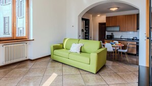 A living room with a green sofa, a dining area with a table and chairs, a kitchen with wooden cabinets, and a window with shutters. - Casa Luvit by Interhome (Menaggio)