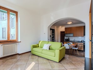 Ein Wohnzimmer mit einem grünen Sofa, ein Essbereich mit Tisch und Stühlen, eine Küche mit Holzschränken und ein Fenster mit Rollläden.