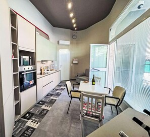 A compact kitchen with built-in appliances, a dining area with chairs, and a large window with blinds. - Il Funghetto by Interhome (Ventimiglia)