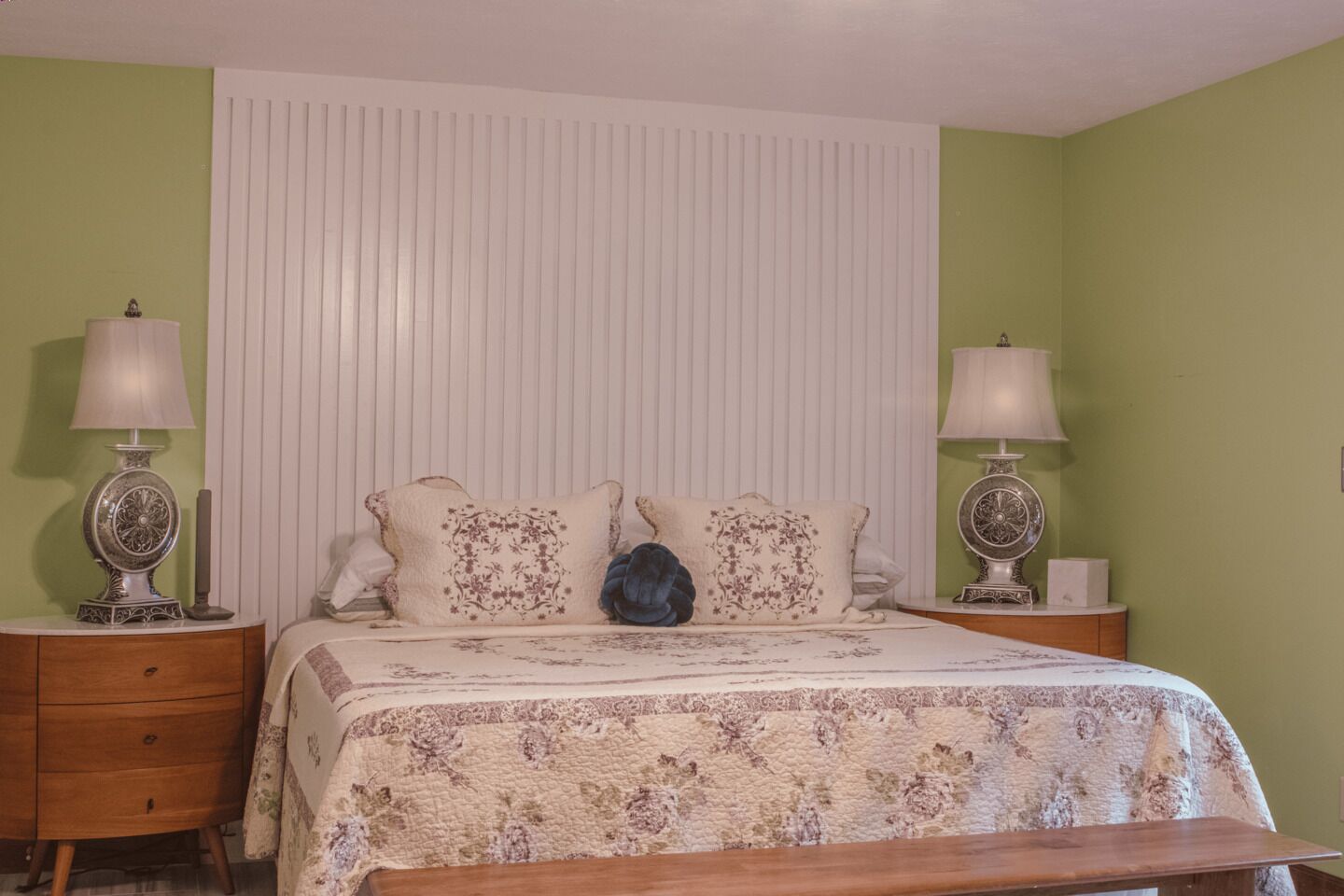A bedroom with a bed, two nightstands, a bench, and a window with blinds.