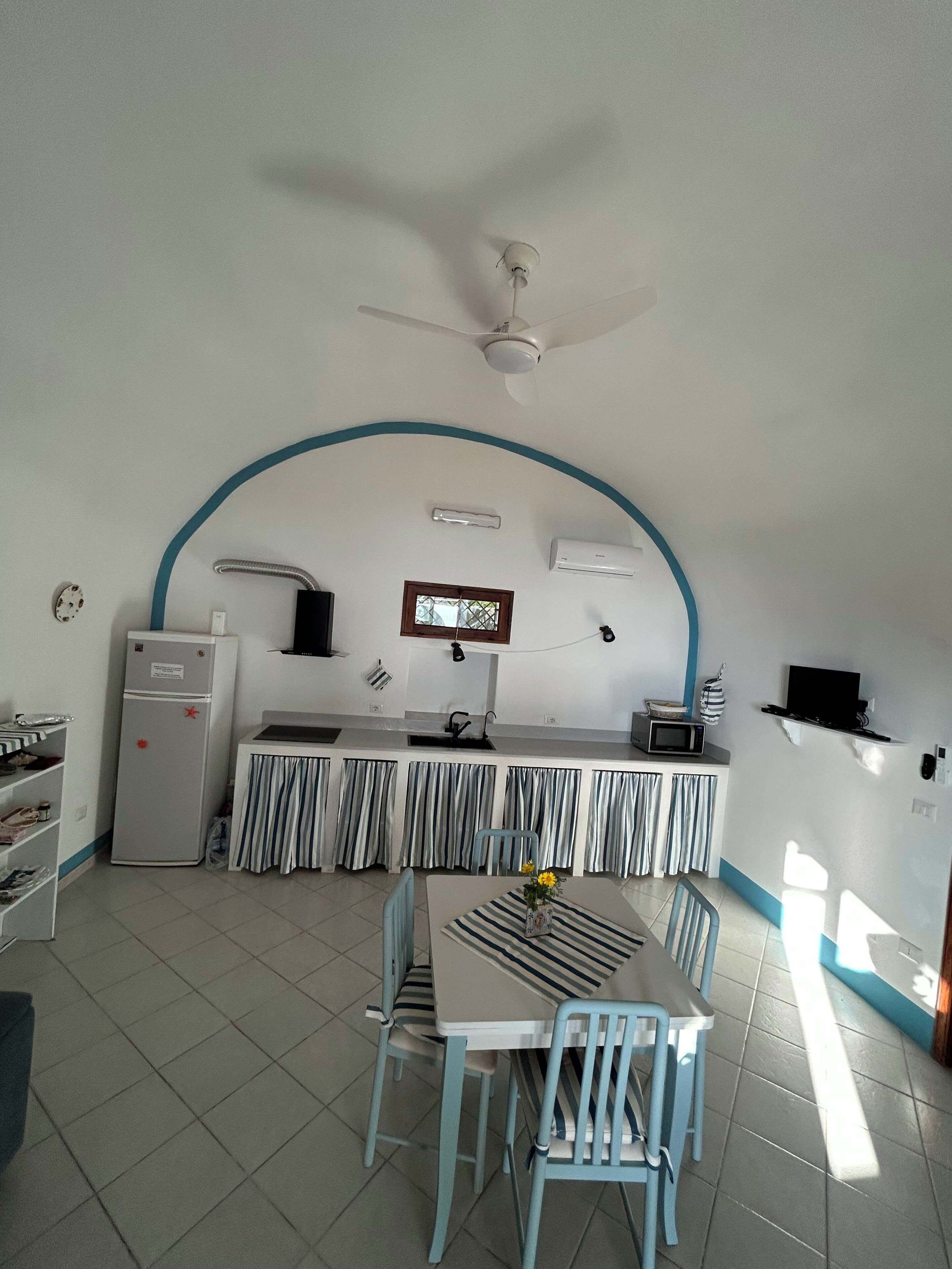 A compact kitchen with a refrigerator, microwave, and stove, a dining area with a table and chairs, and a ceiling fan.