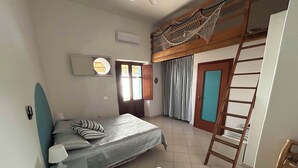 A bedroom with a bunk bed, a single bed, a bedside table, a lamp, a window with curtains, an air conditioning unit, and a wooden ladder. - Melania House II (Filicudi Porto)