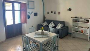 A dining area with a table and chairs, a sofa, a bookshelf, and a door leading outside. - Melania House II (Filicudi Porto)