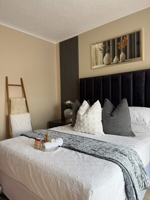 A neatly made bed with a white and gray patterned bedspread, a dark headboard, and a bedside table with a vase of dried flowers. - The perfect 2-bedroom apartment Designed for those who value neatness. (Harare)