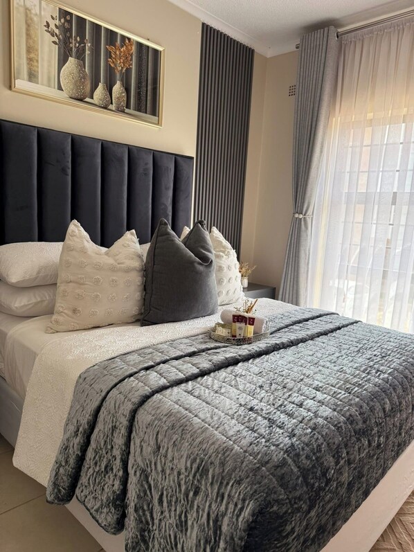 A bedroom with a bed, pillows, a bedside table, a mirror, and a window with curtains.