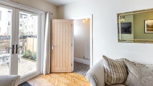 Een kamer met een visgraatparketvloer, een bank met gestreepte kussens, een glazen schuifdeur naar buiten en een lijstbeeldspiegel aan de muur.