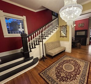A staircase with black and white balusters, a chandelier, a tufted sofa, and a patterned rug. - Historic Belle Maison: Billiards Room, Fire Pit & Magnolia Charm (Magnolia)