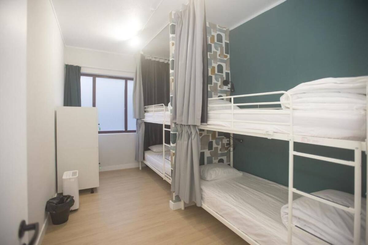 A dormitory room with bunk beds, a window, a white radiator, and a trash bin.