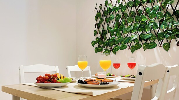 Une table à manger avec des assiettes remplies de nourriture et des boissons, des chaises blanches et une plante fixée au mur.