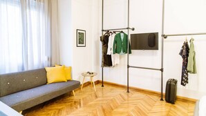 Ein Zimmer mit grauem Sofa, gelben Kissen, einem kleinen runden Tisch, einem Kleiderständer und einem Koffer.