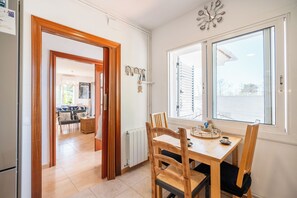 Un coin repas avec une table et des chaises en bois, une fenêtre avec des stores, un radiateur et une porte donnant sur une autre pièce.