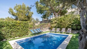 Une piscine dont le bord est recouvert de carreaux, entourée de pelouse verdoyante et d’arbres, avec des chaises longues et un mur de pierre en arrière-plan.