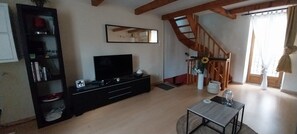 Una sala de estar con televisor, estantería, escalera y ventana con cortinas.