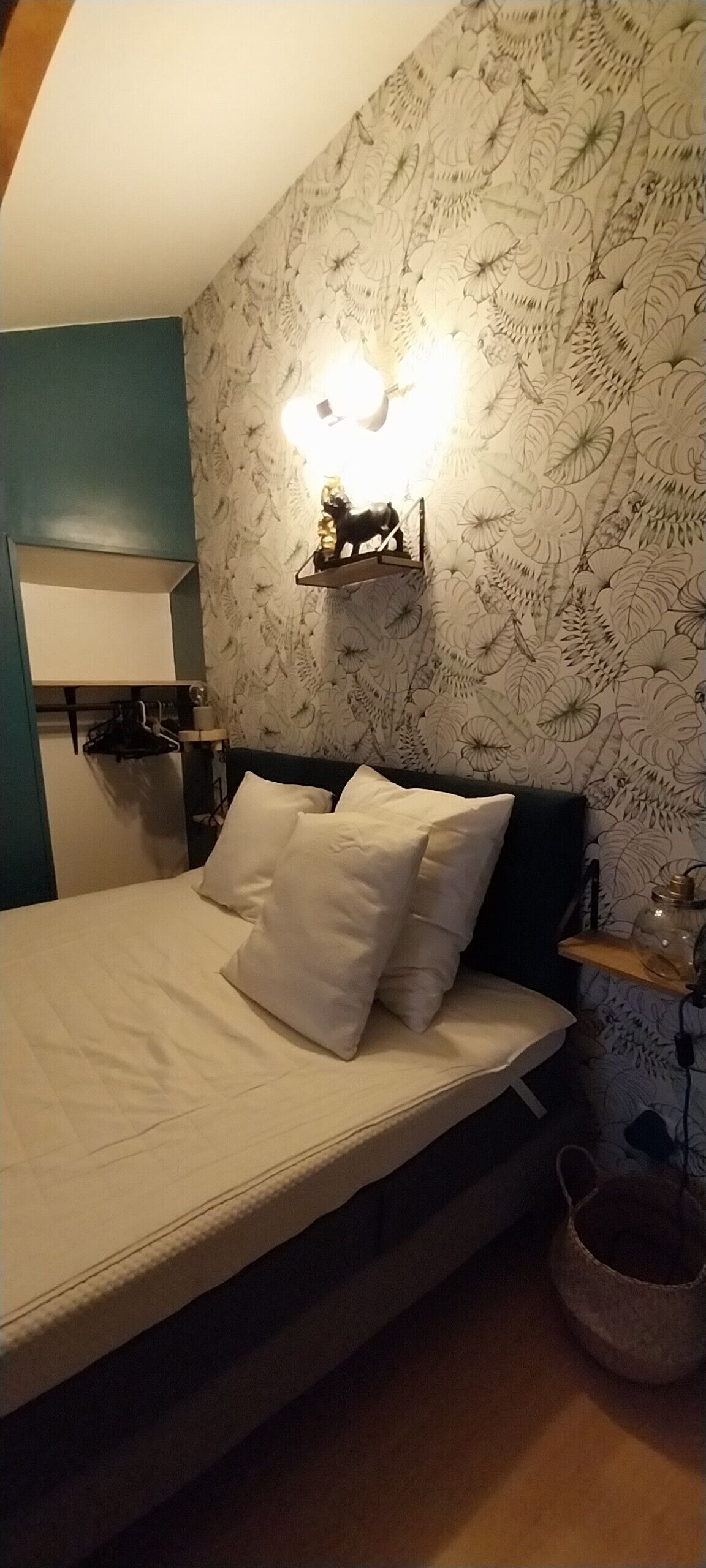 A bedroom with a bed, pillows, a nightstand, a lamp, and a wall with a floral pattern.