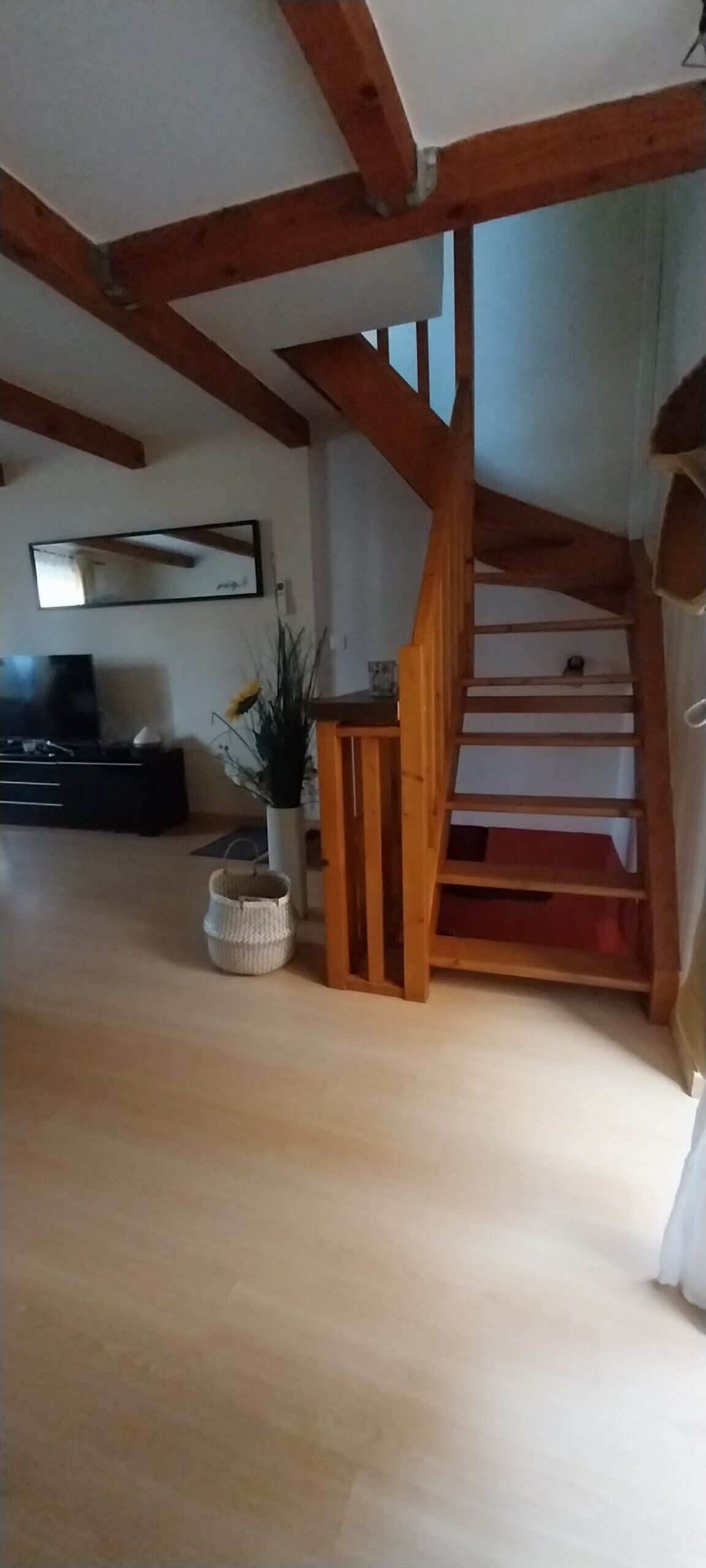 Ein Loft-Zimmer mit freiliegenden Holzbalken, einer Holztreppe, einem Flachbildfernseher, einem Topf mit Pflanze und einem gewebten Korb.