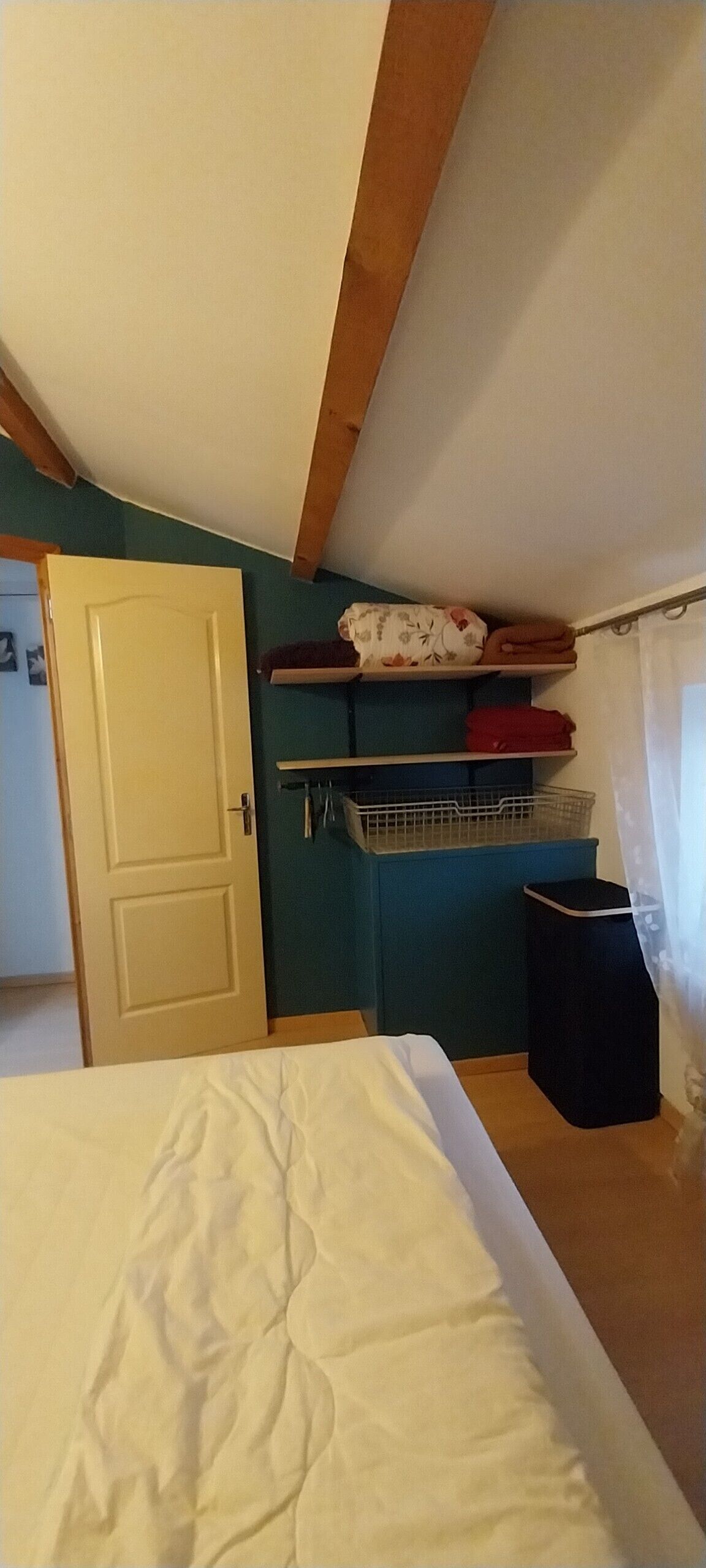 A bedroom with a bed, a door, a shelf with items, and a window with curtains.