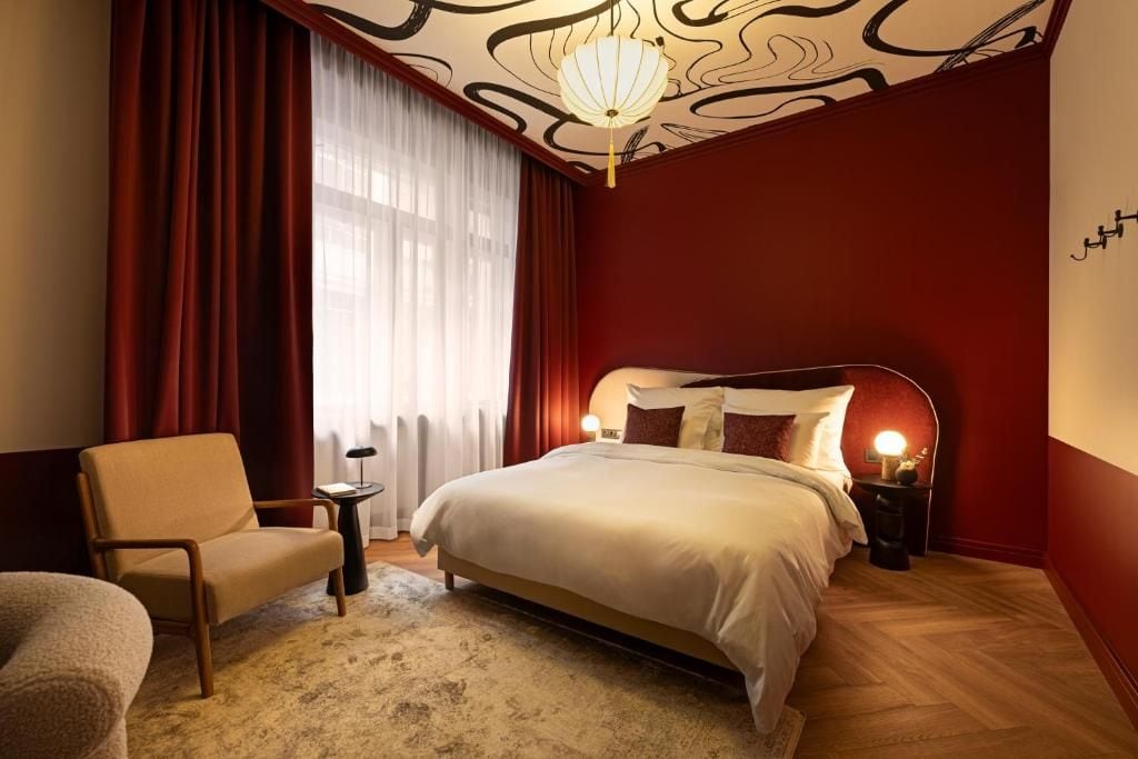 A bedroom with a bed, a chair, a nightstand, a lamp, and a window with curtains.