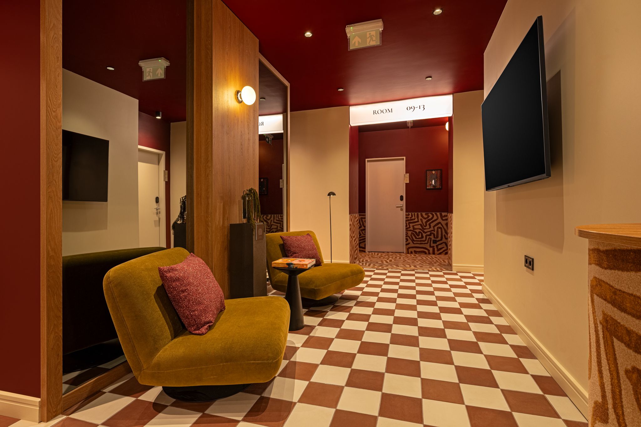 Un hall de hotel con piso a cuadros, dos sillones con cojines, un televisor de pantalla plana en la pared y una puerta con la leyenda 'ROOM 09-13'.