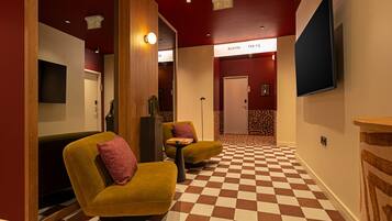 Un hall de hotel con piso a cuadros, dos sillones con cojines, un televisor de pantalla plana en la pared y una puerta con la leyenda 'ROOM 09-13'.