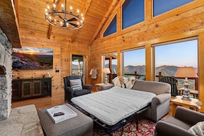 Interior de cabana de tronco com cama, área de estar e grandes janelas com vista para as montanhas.