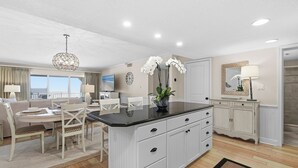 A modern kitchen with a central island, white cabinetry, and a dining area with a table and chairs. - 🌊Beachfront Stunner | Panoramic Ocean Views | Great Amenities | Sandpiper 514🌊 (New Smyrna Beach)
