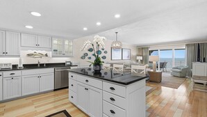 A modern kitchen with white cabinetry, a central island, and a dining area with a view. - 🌊Beachfront Stunner | Panoramic Ocean Views | Great Amenities | Sandpiper 514🌊 (New Smyrna Beach)