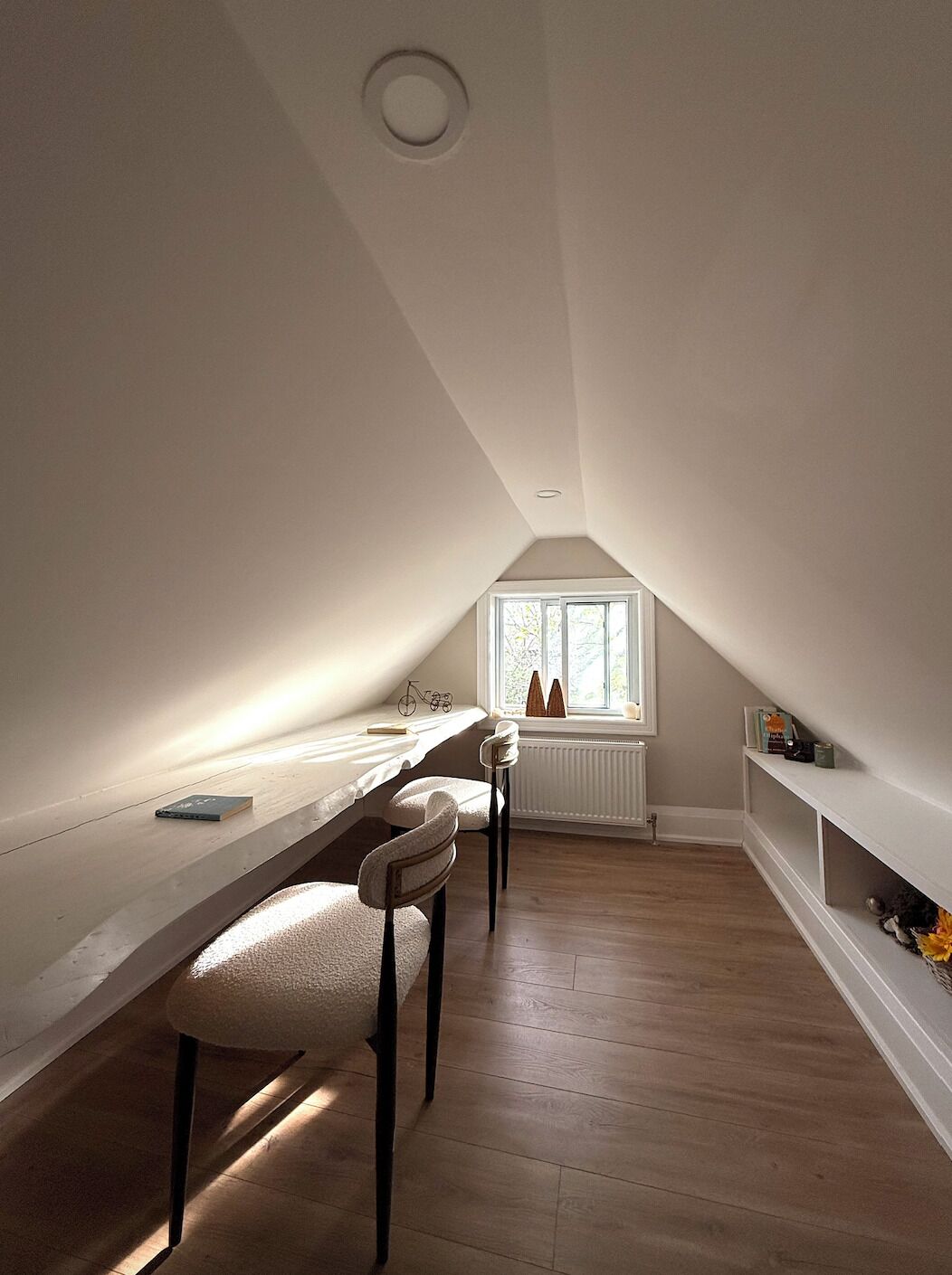 A room with a sloped ceiling, a window with a view, a radiator, and a long table with chairs.