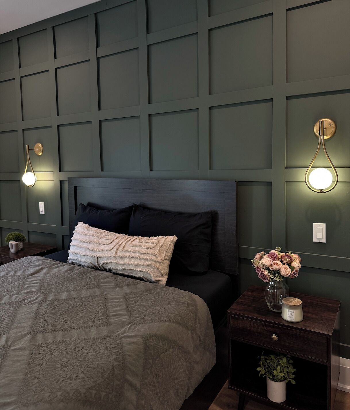 A bedroom with a bed, dark headboard, two bedside tables with lamps, a nightstand with a vase of flowers, and a wall panel design.