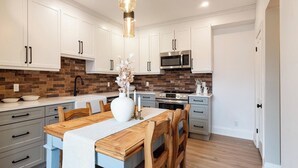 A modern kitchen with white cabinets, a wooden dining table, and a brick backsplash. - Muskoka Stay in Downtown Huntsville with Firepit (Huntsville)