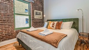 Ein Zimmer mit Backsteinwand, großem Fenster, Bett mit grünem Kopfteil und Nachttisch.