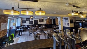 A restaurant interior with multiple tables, hanging pendant lights, and a bar area with taps. - Les Marais Comtois (Saône)