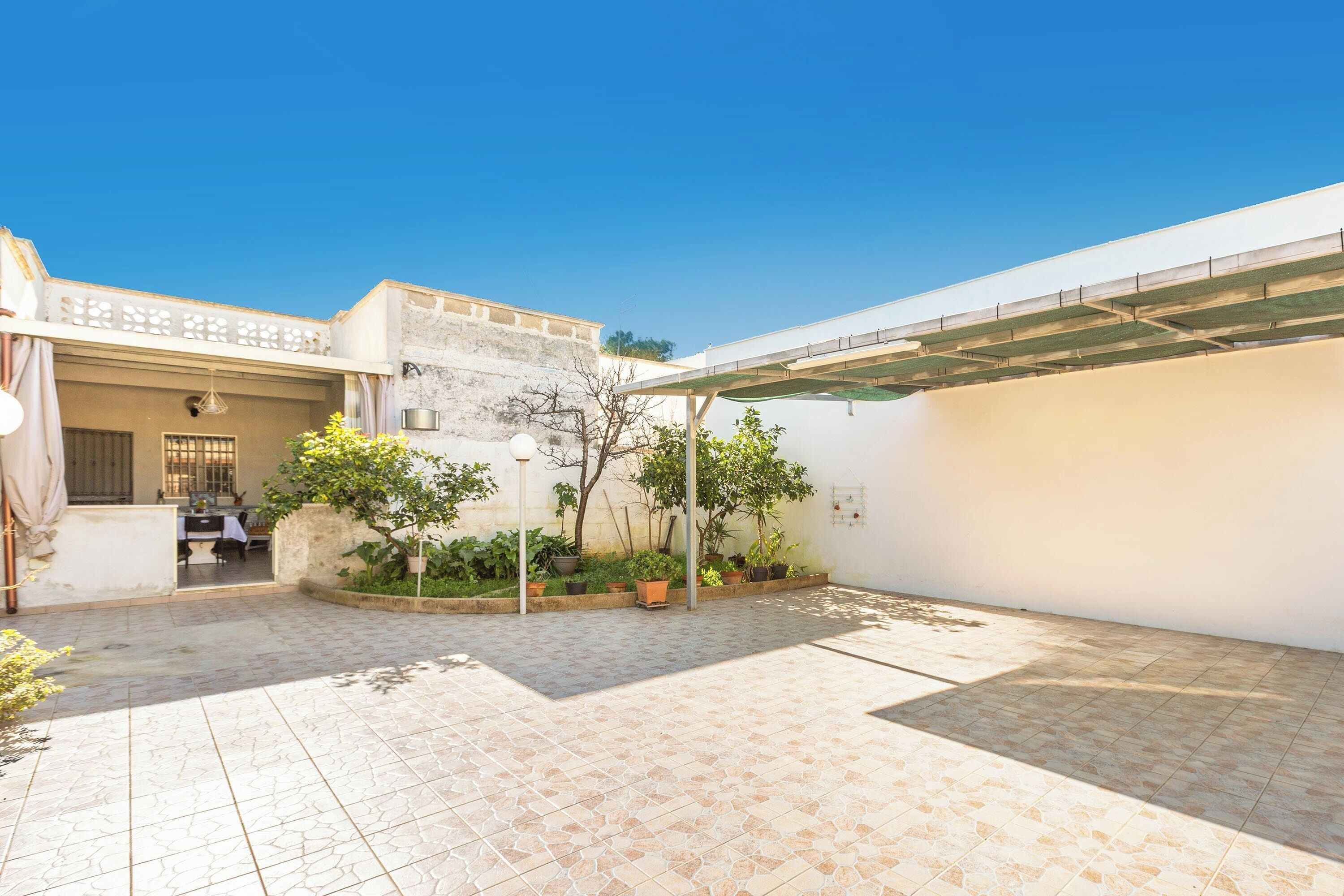 Um pátio com piso de azulejos, plantas em vasos, uma pérgola e um edifício com janela e porta.