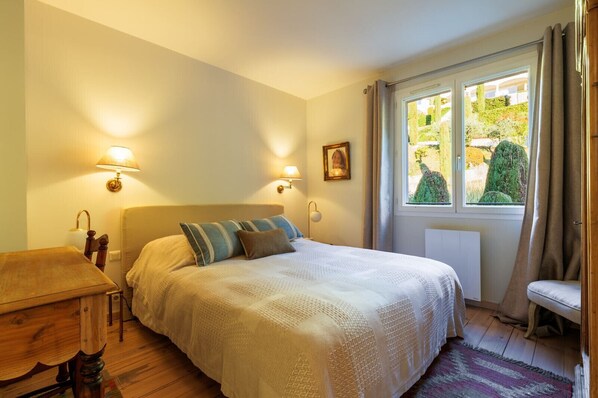 Ein Schlafzimmer mit einem Bett, Nachttischlampen, einem Fenster mit Vorhängen, einer Heizung und einem Holztisch.