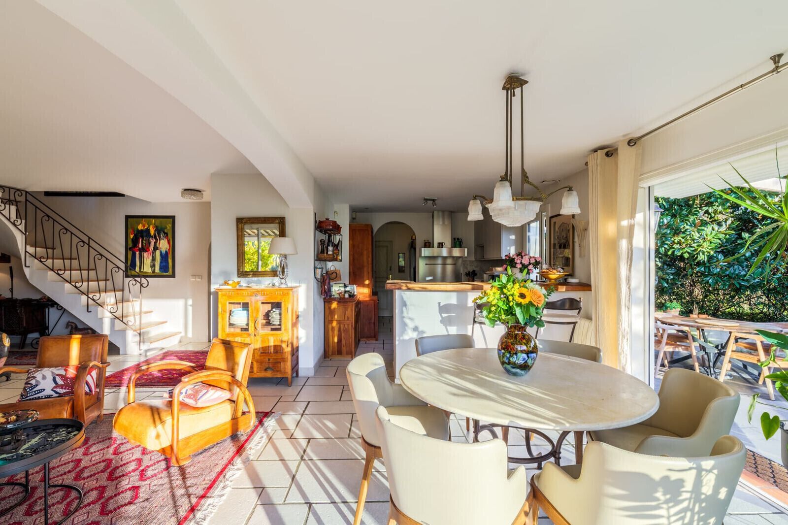 A spacious dining area with a round table and chairs, a kitchen in the background, and a staircase leading to another level.