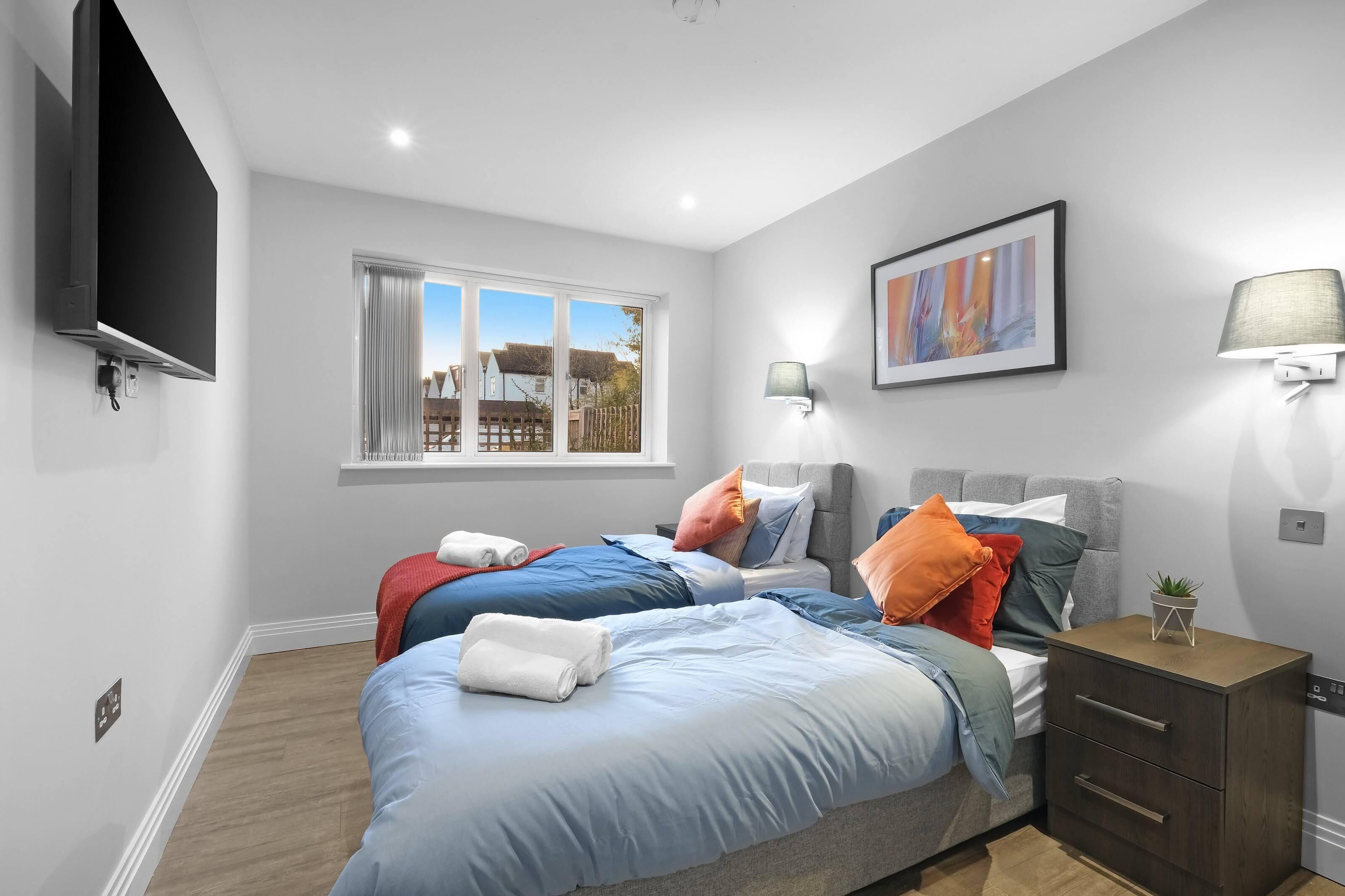 A bedroom with two beds, a TV, a window, a painting, and a bedside lamp.