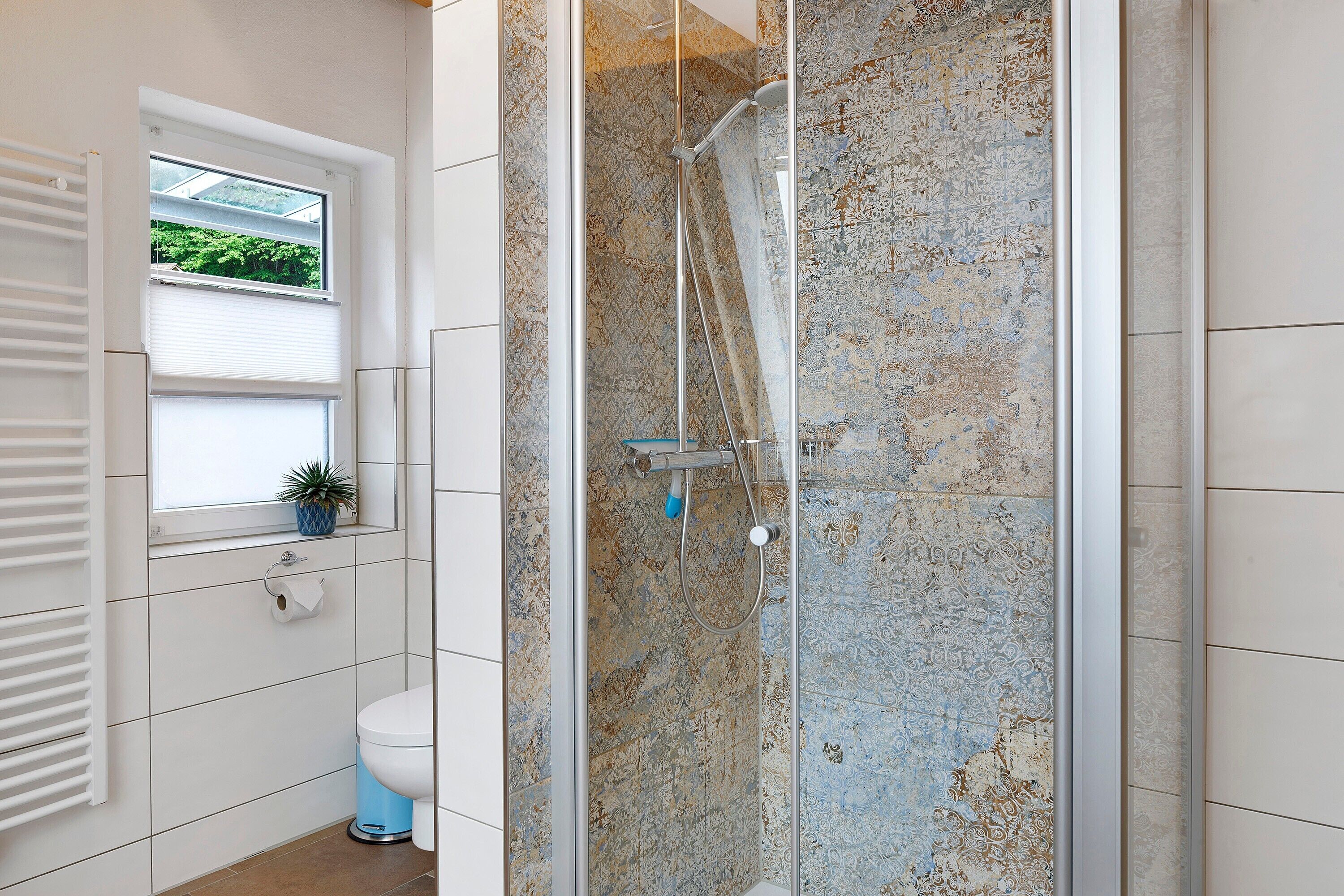 Ein Badezimmer mit Duschkabine, Toilette, Fenster mit Jalousien und einem kleinen Topf auf der Fensterbank.