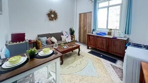 Un salon comprenant un canapé, une table basse et un coin repas avec une table et des chaises. Il y a une fenêtre avec des rideaux, un ventilateur de plafond et une porte.