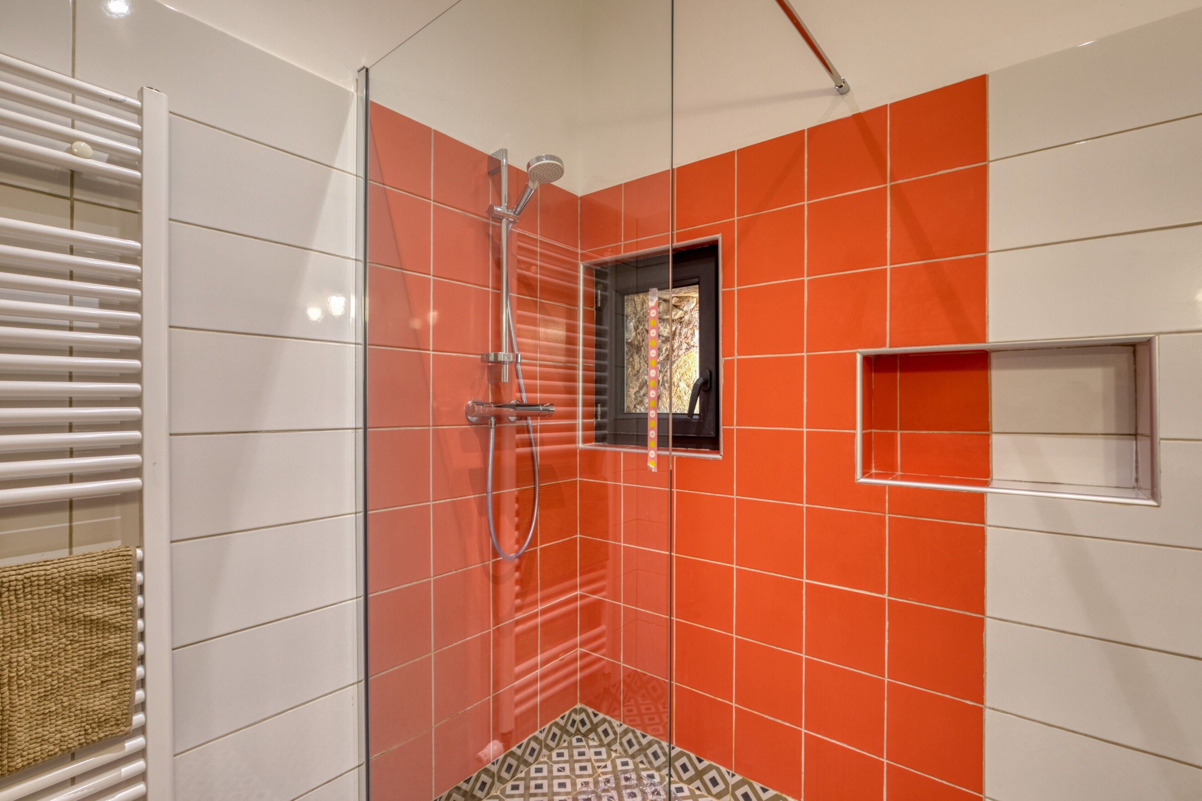 A shower with a glass door, a towel rack, and a small window with a colorful pattern.