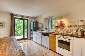 A kitchen with a wooden table, a refrigerator, a dishwasher, a stove, and a window with a view of greenery. - Holiday Home “Le Moulin de Mirande” with Terrace, Private Garden & Wi-Fi (La Tour-sur-Orb)