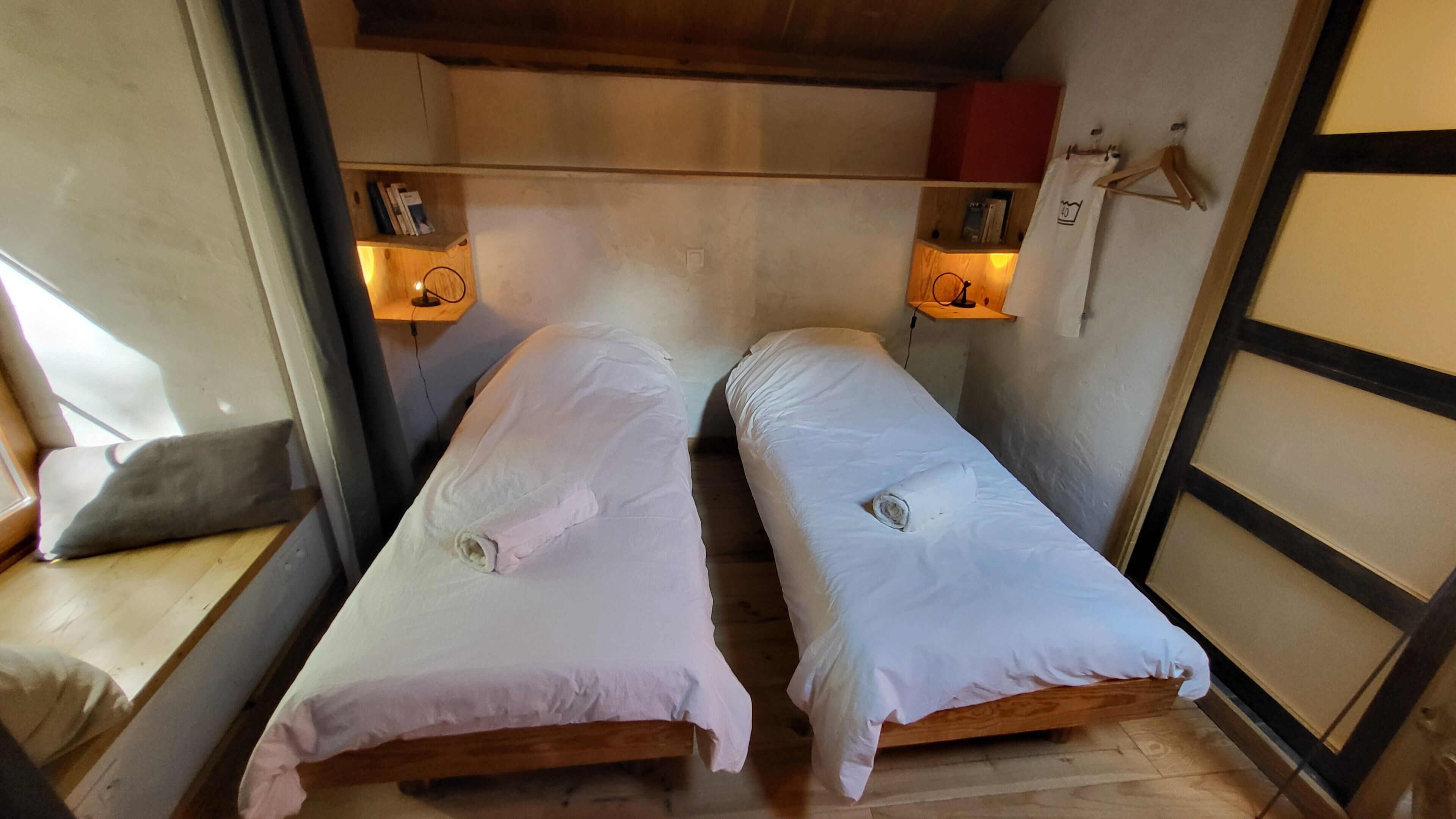 Een knus, compacte kamer met twee eenpersoonsbedden, een houten bankje en een klein plankje met een lamp.